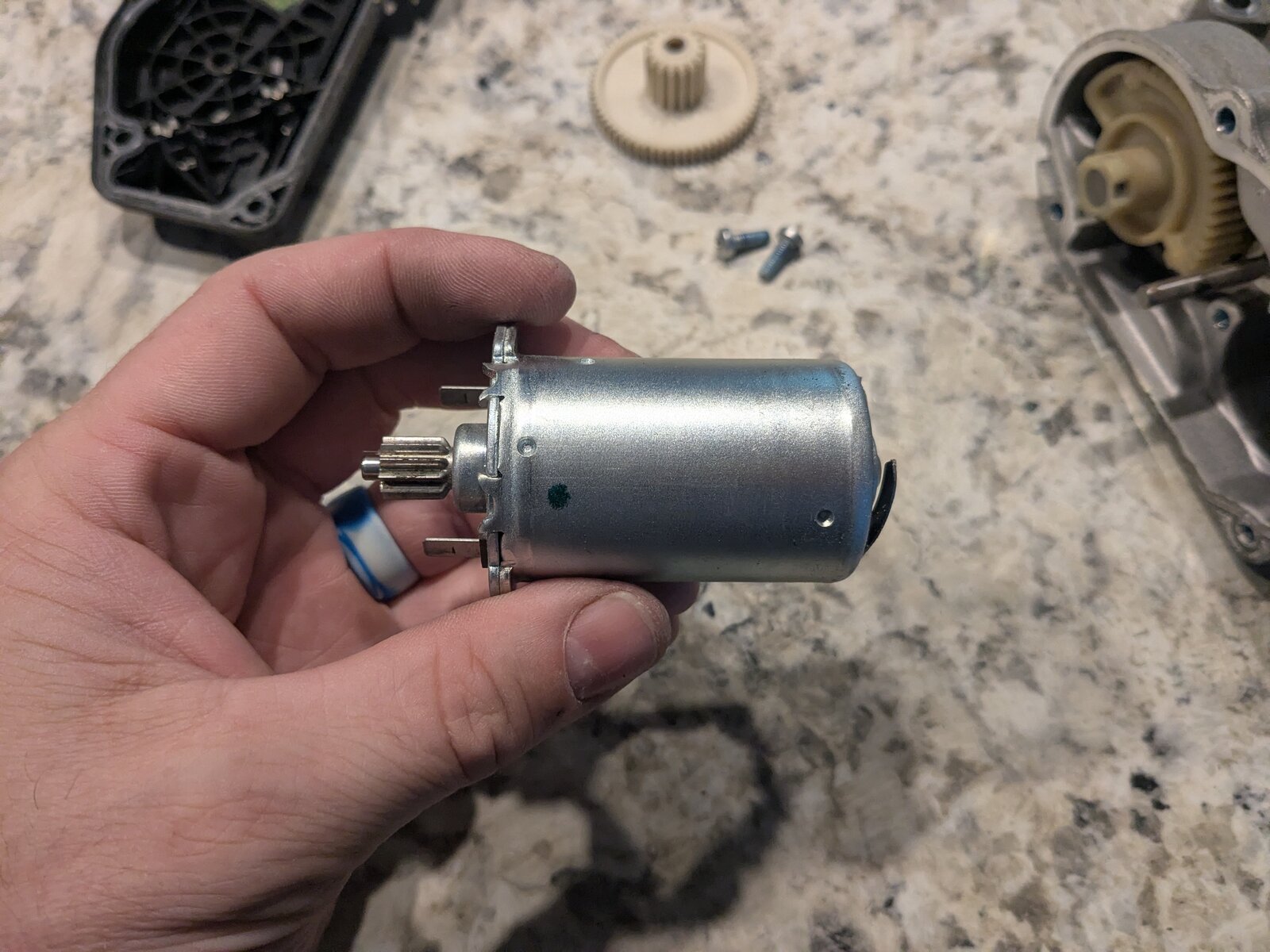 The small DC actuator motor removed from the housing, held in hand, showing its output pinion gear and two spade electrical terminals.