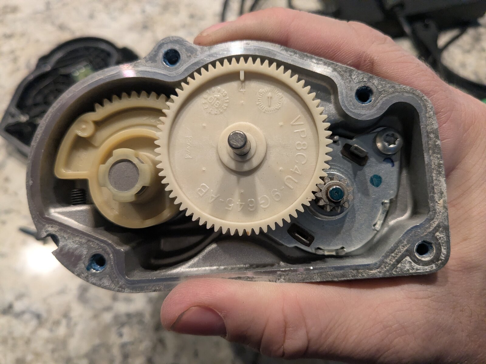 Closer view of the final output gear, stamped VP8C4U-9GG45-XB, meshed with the smaller motor pinion gear and a spring-loaded return mechanism.